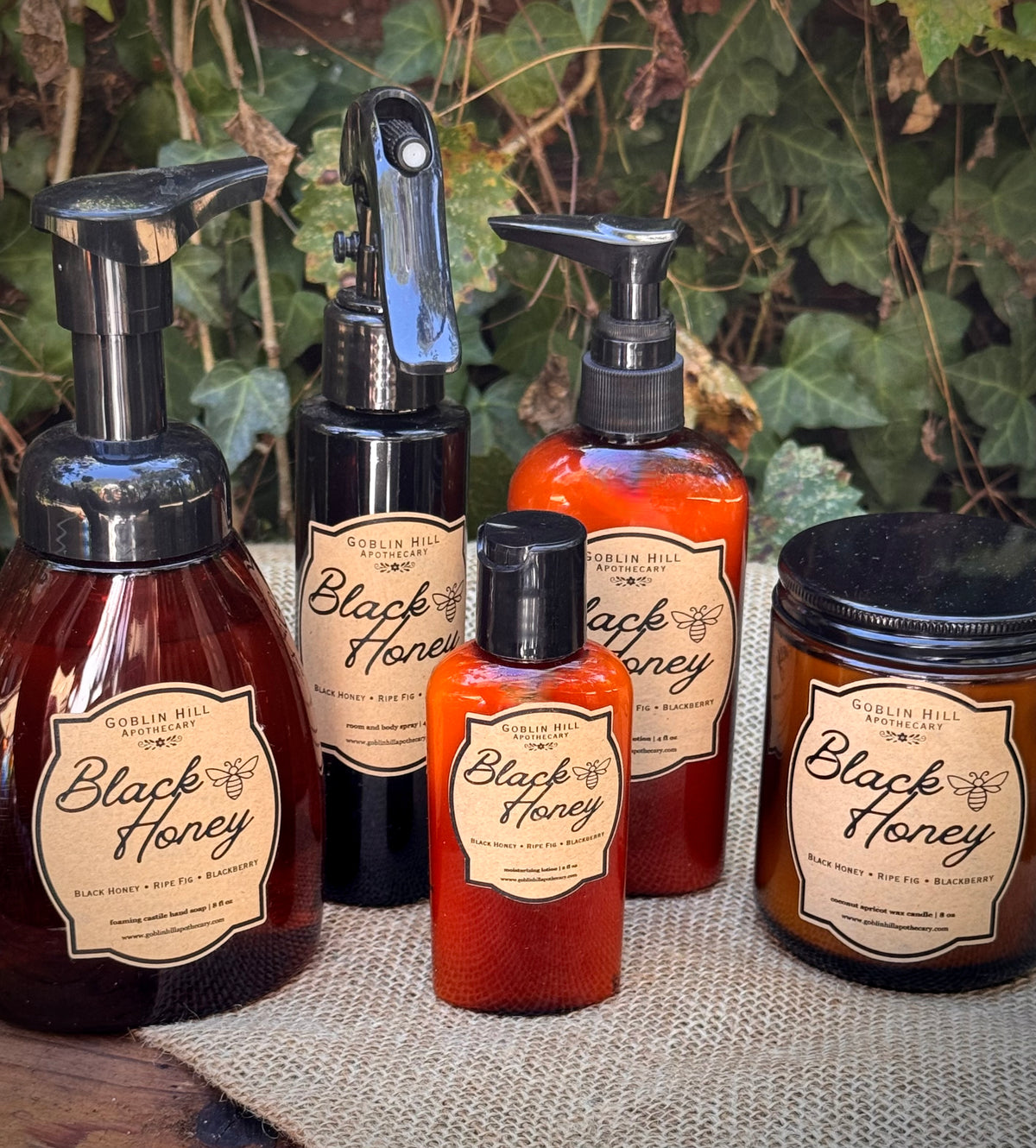 Five Goblin Hill 'Black Honey' products arranged on a burlap surface with ivy in the background: a large amber pump bottle, a black spray bottle, a red-orange pump bottle, a small red-orange bottle, and a dark amber jar.