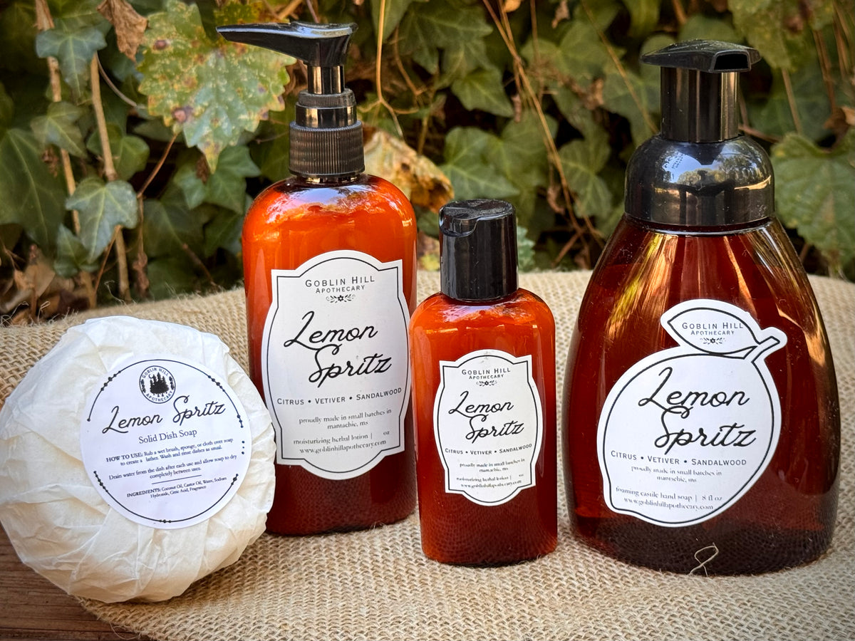 A set of Lemon Spritz hand and body care products from Berlin Hill Apothecary, including a wrapped bar of soap, a pump bottle, a small bottle, and a foaming dispenser, arranged on a burlap surface with ivy in the background.