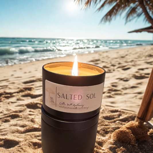Candle labeled 'Salted Sol' on a sandy beach with ocean and palm tree in the background