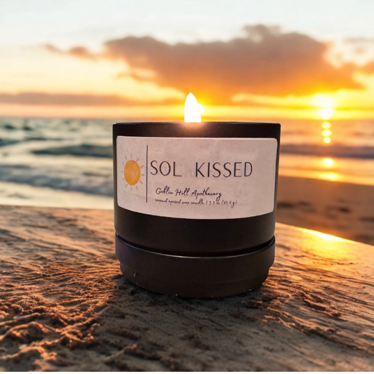 Candle labeled 'SOL KISSED' on a beach at sunset