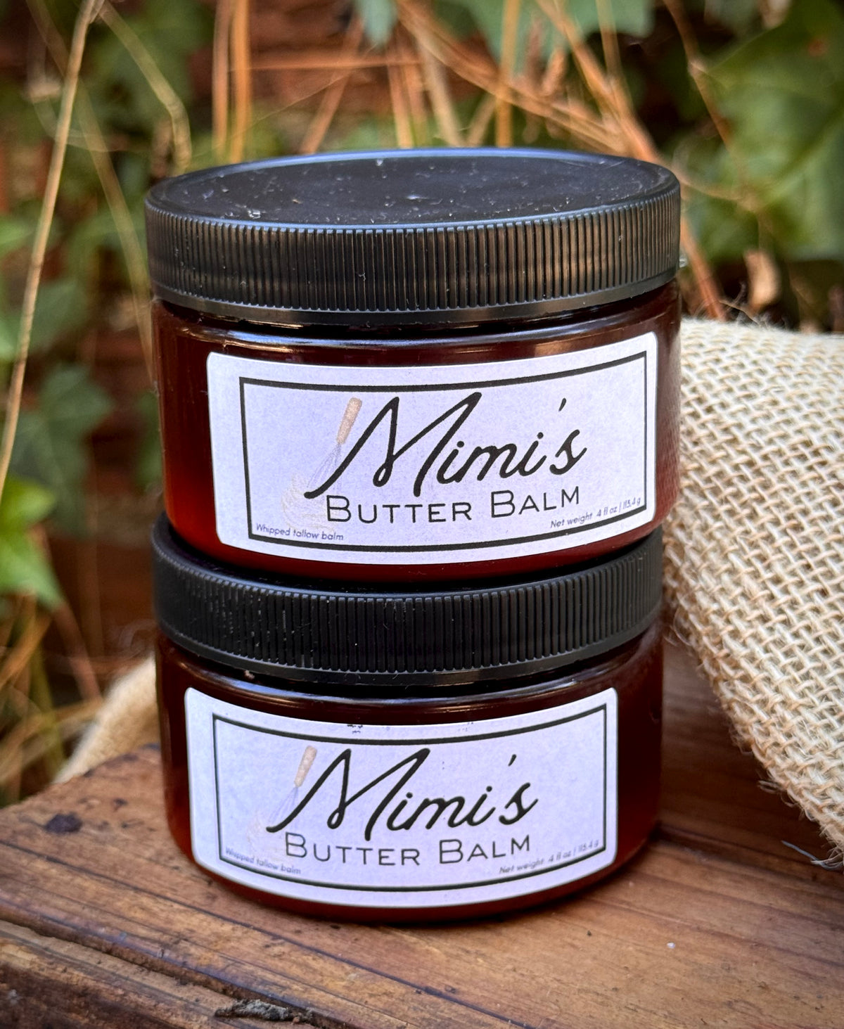 Two stacked amber glass jars of Mimis Butter Balm with black lids and white labels, set on a rustic wooden surface with burlap and green foliage in the background.