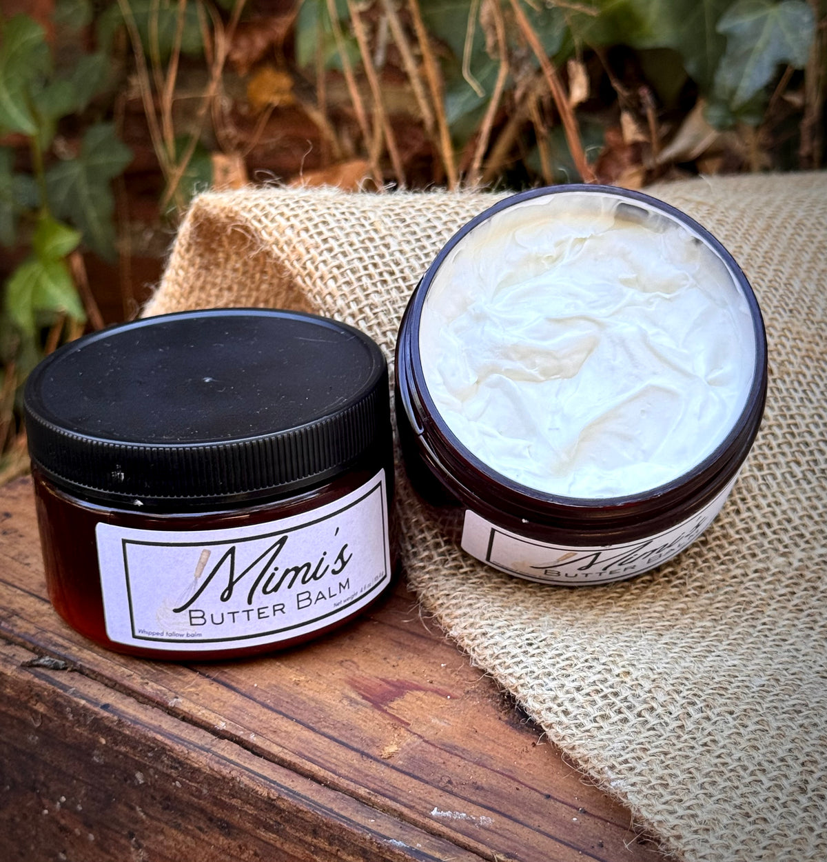 Two jars of Mom's Butter Balm displayed on a rustic wooden surface with burlap and green foliage in the background. One jar is closed with a black lid, and the other is open to show the thick white butter balm inside.
