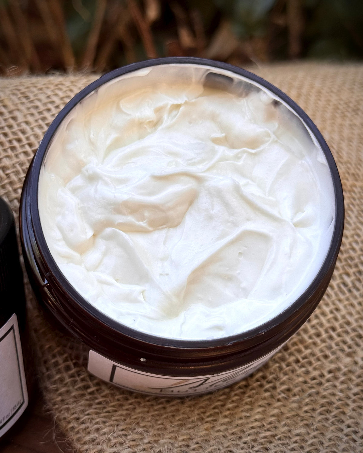An open dark amber jar filled with thick white whipped cream or balm, placed on a textured tan burlap surface with a blurred natural background.
