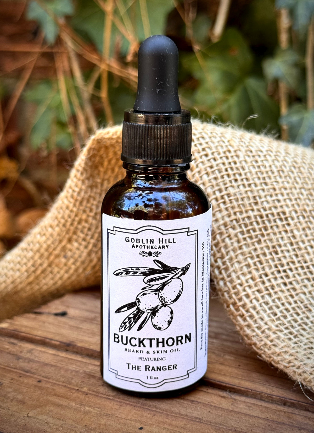 A 10ml amber glass dropper bottle of Buckthorn Beard & Skin Oil by Goblin Hill Apothecary, set on a rustic wooden surface with a burlap cloth and blurred green foliage in the background.