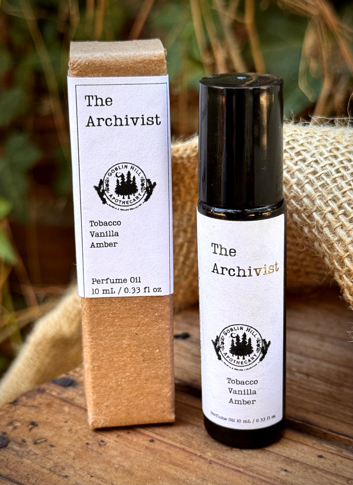 A bottle of 'The Archivist' Tobacco Vanilla Amber perfume oil stands next to its kraft paper packaging box on a rustic wooden surface with burlap and green foliage in the background.