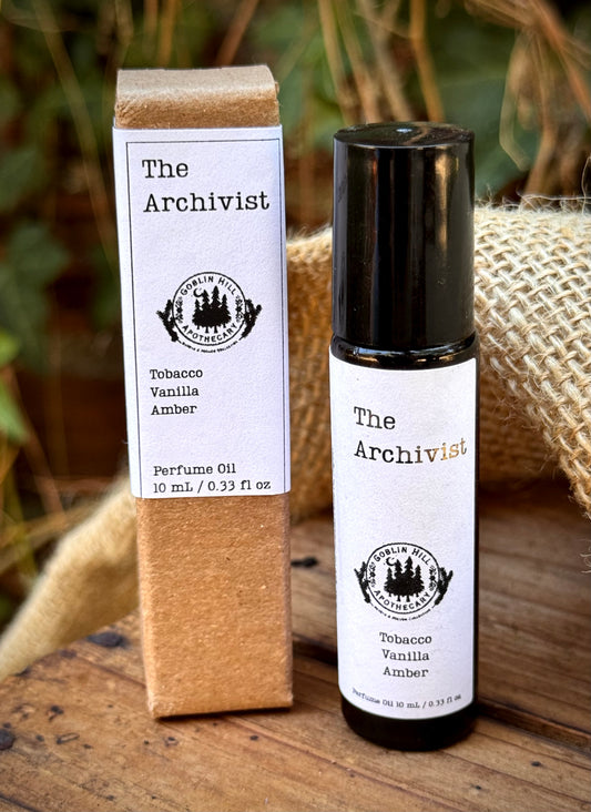 A bottle of 'The Archivist' Tobacco Vanilla Amber perfume oil stands next to its kraft paper packaging box on a rustic wooden surface with burlap and green foliage in the background.