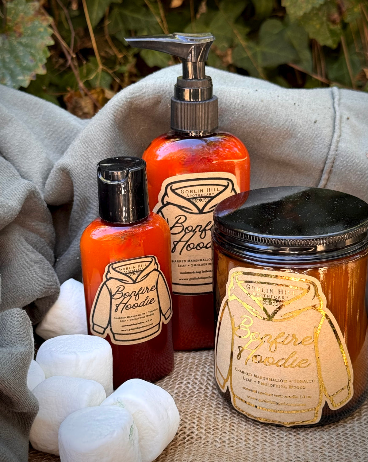 Three Borflame Hoodie scented products arranged on burlap with a gray hoodie and marshmallows in the background: a pump bottle, a smaller bottle, and a candle jar, all with cream labels featuring a hooded ghost outline.