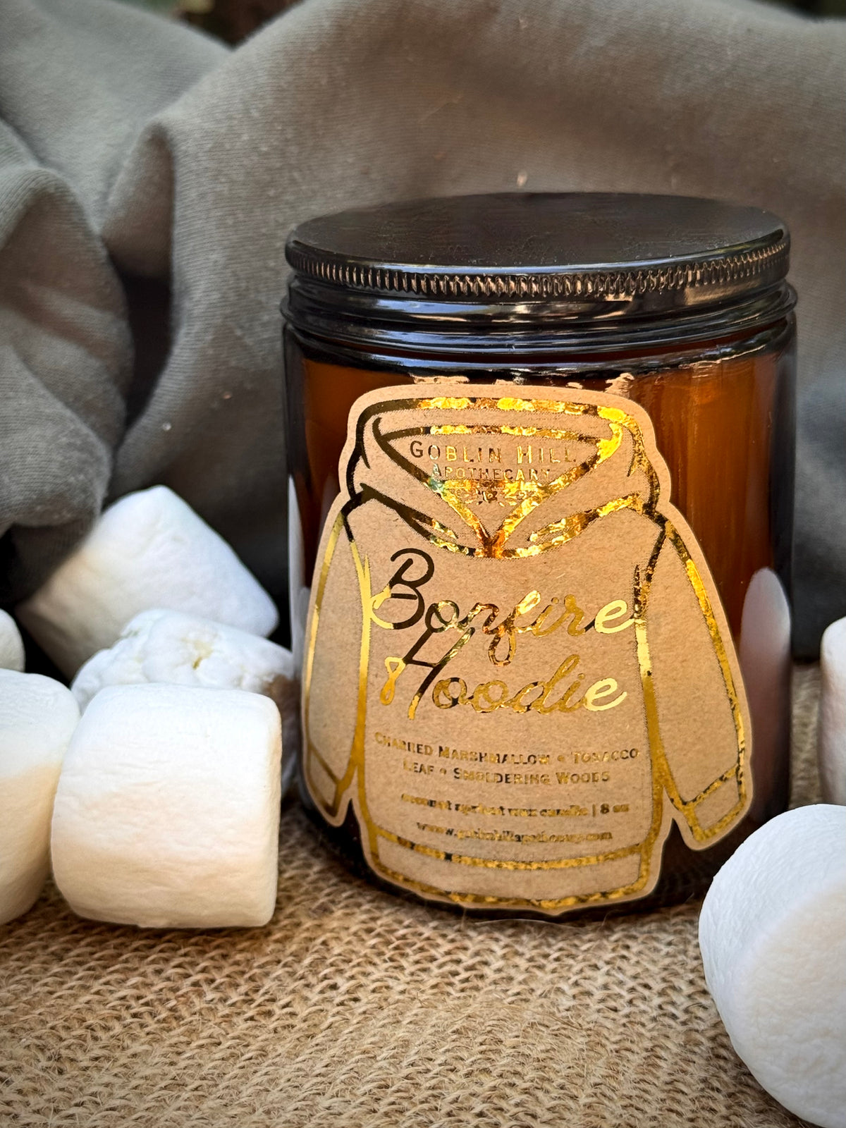An amber glass jar candle with a black lid and a gold foil hoodie outline label reading 'Bonfire Hoodie,' styled with marshmallows on burlap against a soft gray fabric background.