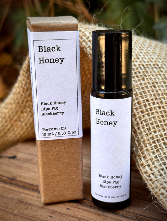 A bottle of Black Honey perfume oil with a white label and black cap stands next to its brown kraft paper box on a rustic wooden surface with burlap in the background.