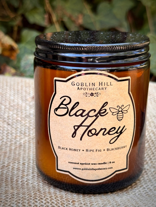 An amber glass jar candle with a black lid and a beige label reading 'Goblin Hill Apothecary Black Honey,' placed on a burlap surface with blurred greenery in the background.