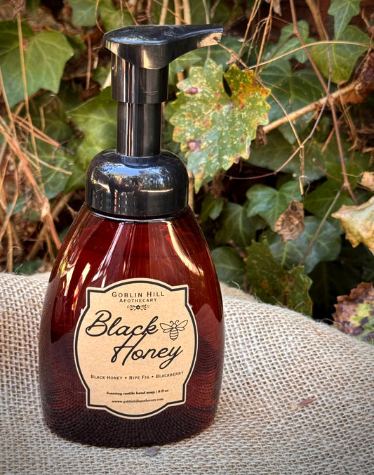 A bottle of Goblin Hill  Black Honey hand wash with a black pump dispenser, set on a burlap sack with green ivy foliage in the background.