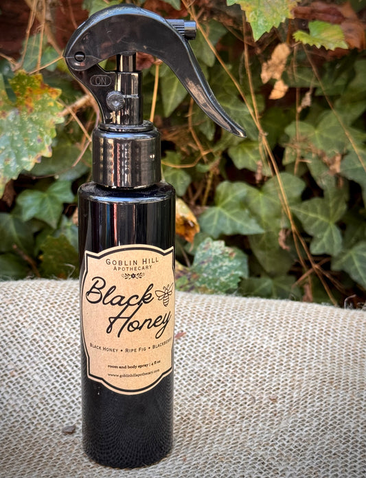 A black spray bottle of Goblin Hill Apothecary Black Honey body spray and perfume oil, placed on a burlap surface with green ivy in the background.