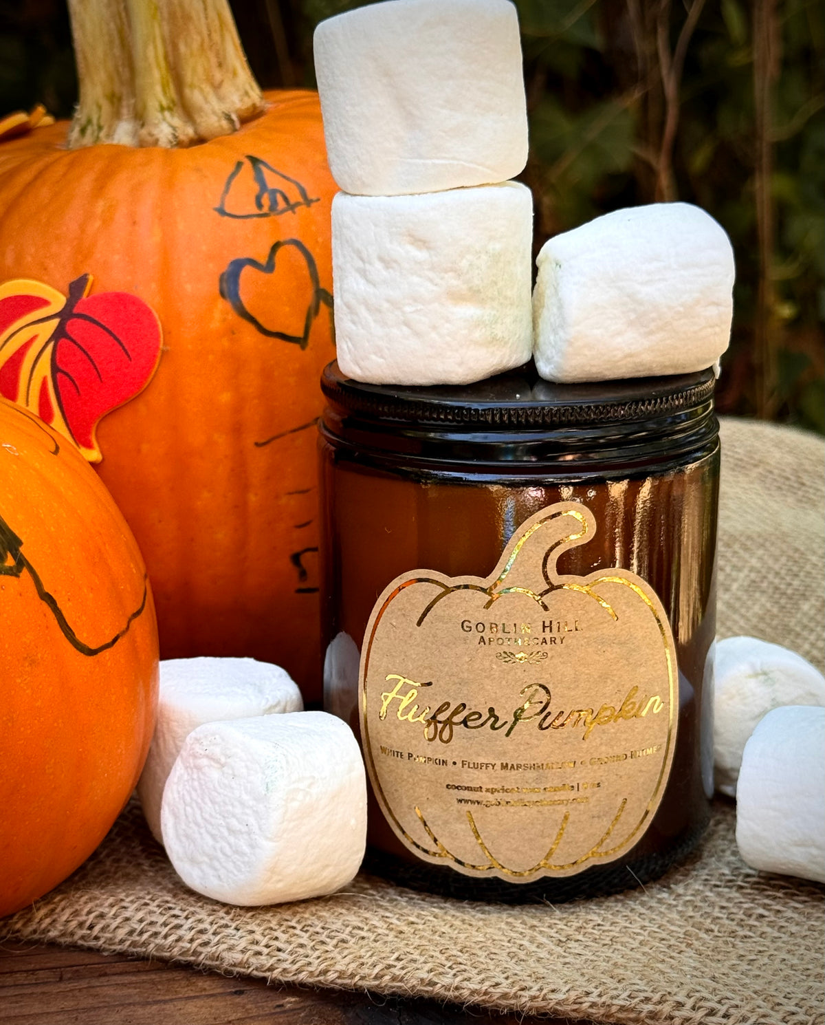 A dark amber glass jar candle with a black lid and a decorative gold label reading 'Hopper Pumpkin' by Goodell Mill Candles, styled with white marshmallows on a burlap surface with pumpkins in the background.
