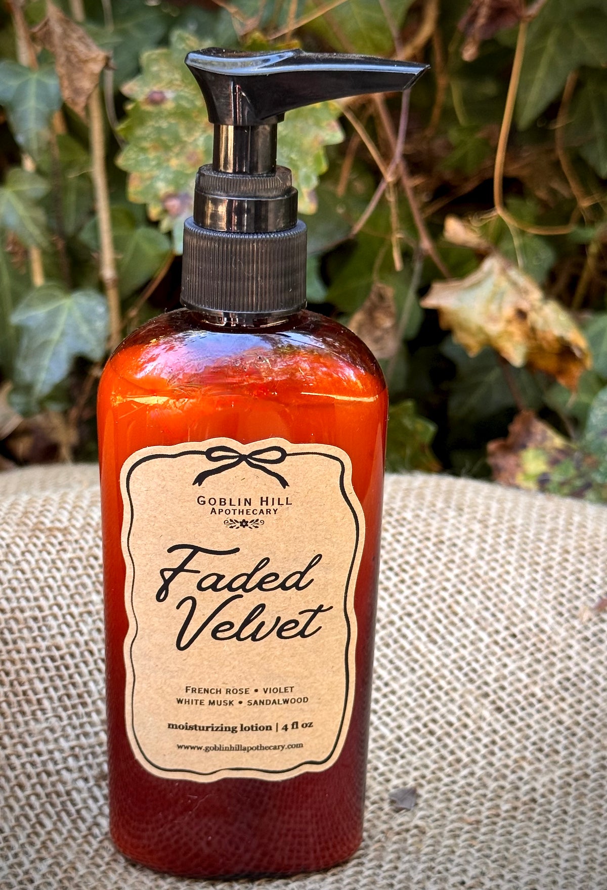 A bottle of Goblin Hill Apothecary 'Faded Velvet' moisturizing lotion, featuring a textured amber-brown bottle and a black pump dispenser, set on a burlap surface with a blurred natural background.