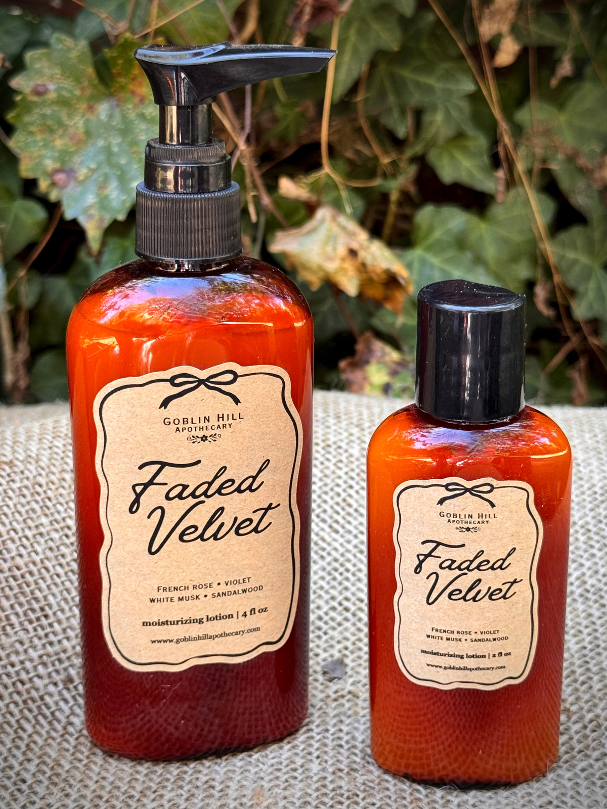 Two Goblin Hill Apothecary Faded Velvet moisturizing lotion bottles, one larger pump bottle and one smaller cap bottle, filled with orange-red liquid and resting on a burlap surface with green foliage in the background.