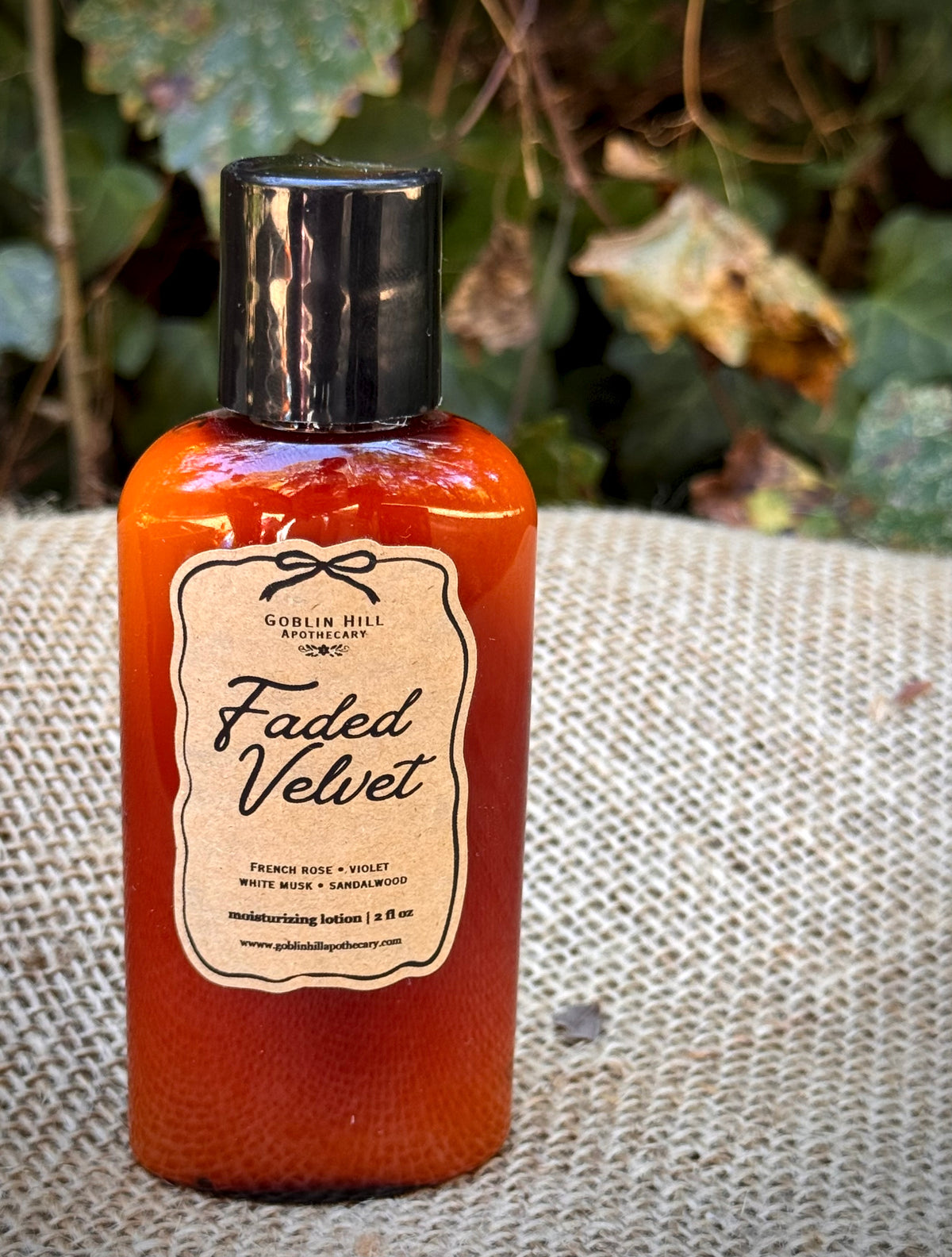 A bottle of Goblin Hill Apothecary 'Faded Velvet' moisturizing lotion, filled with a translucent orange-red liquid, resting on a burlap sack with a blurred natural background.