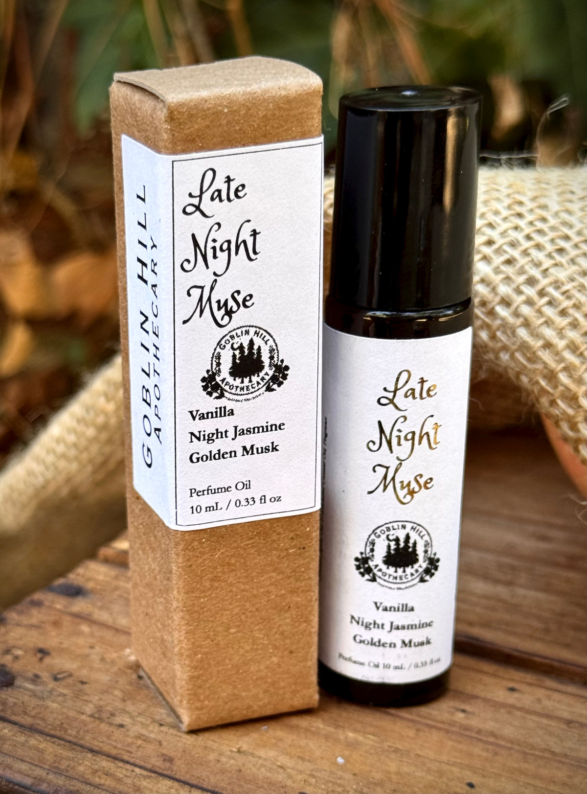 A bottle of perfume oil and its kraft paper packaging box, both labeled 'Late Night Muse' and 'Vanilla Night Jasmine Golden Musk', displayed on a rustic wooden surface with burlap and foliage in the background.