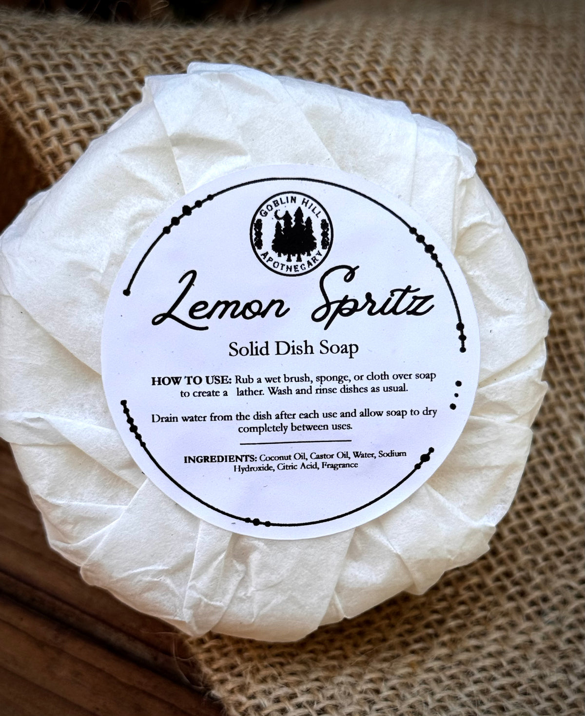 A round bar of Lemon Spritz solid dish soap wrapped in white paper, featuring a white paper label with black text and a circular logo, set on a burlap background.
