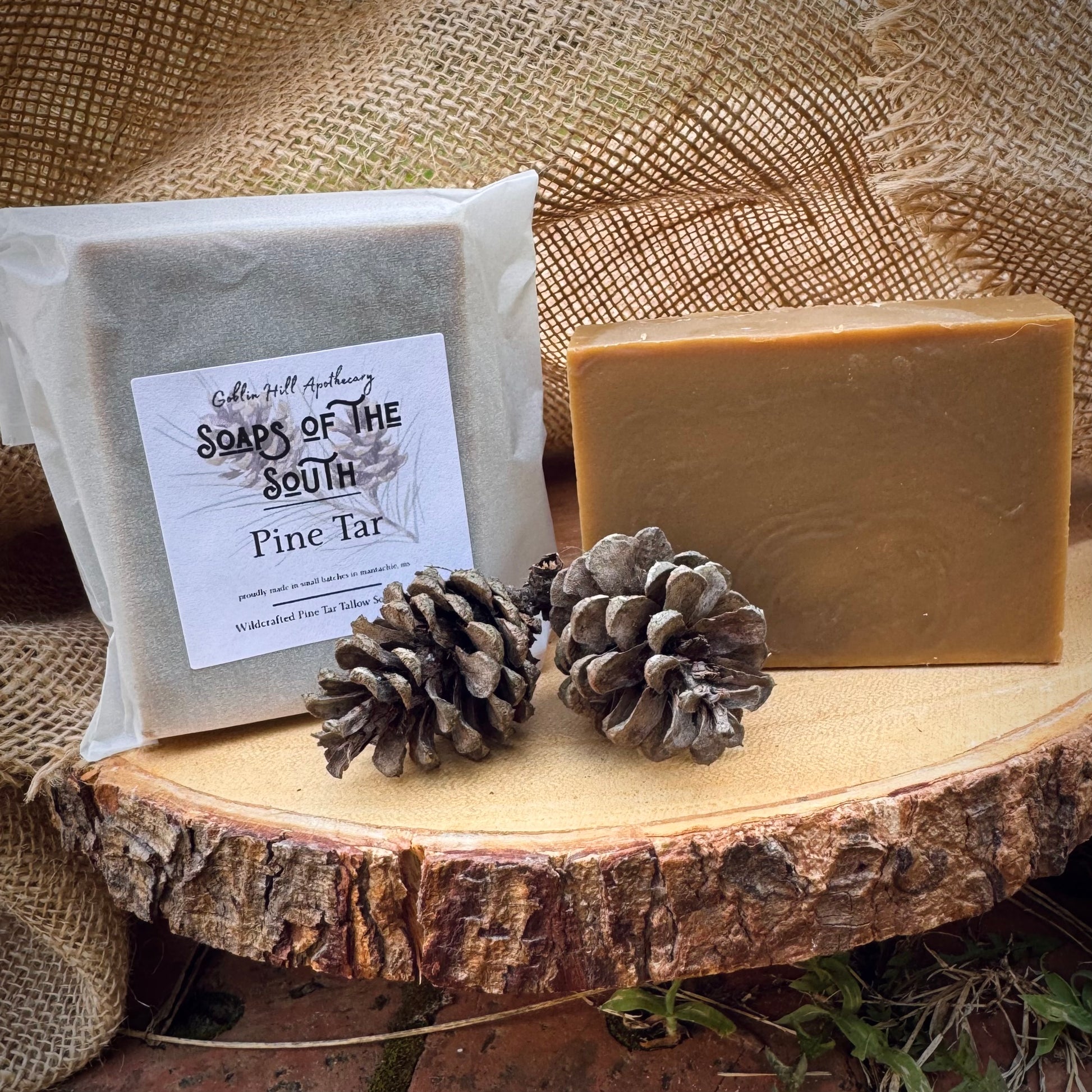 Two bars of soap with a label, two pine cones on a wooden surface with burlap in the background.