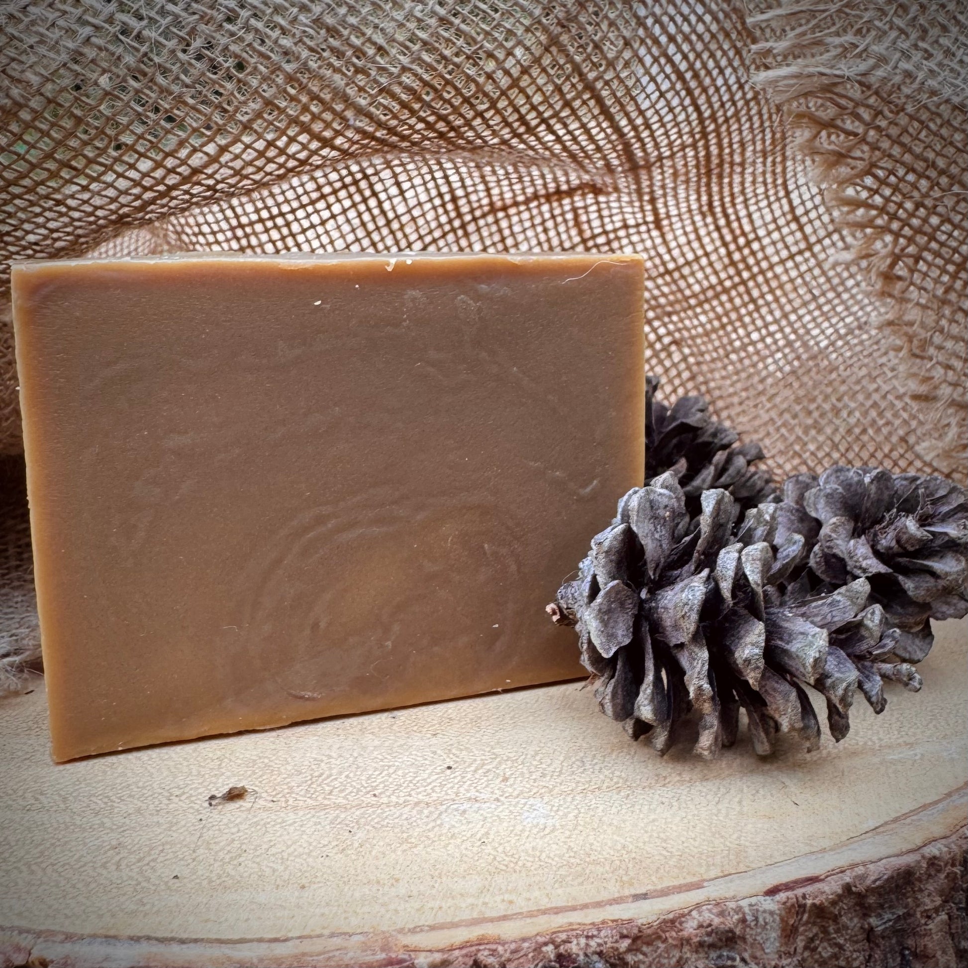 Bar of soap next to a pine cone on a wooden surface with burlap in the background